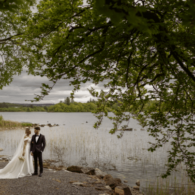 A newlywed couple stands by a tranquil lakeside under lush trees, offering a romantic escape at our hotel retreat.