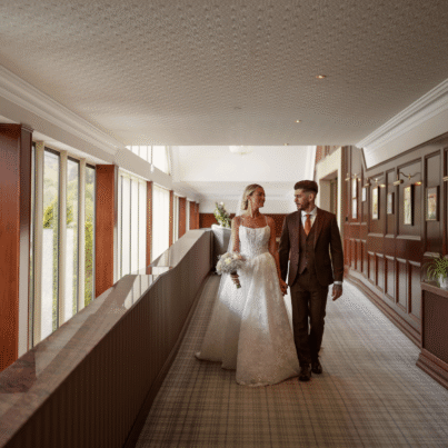 Newlyweds stroll in elegant hotel corridor with scenic views and rich wood panelling.