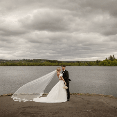 Romantic couple in elegant wedding attire embrace by serene lakeside under cloudy sky, offering stunning rural views.