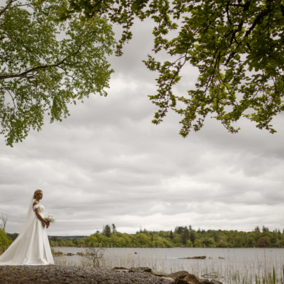 Bride in elegant gown by serene lakeside with lush trees and tranquil water, perfect for romantic weddings and nature retreats.