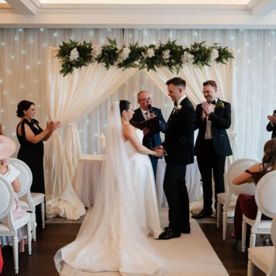 Elegant wedding ceremony in hotel ballroom with floral arch, fairy lights, and seated guests in stylish attire.