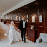Elegant couple walking in refined hotel corridor with wood panelling, soft lighting, and art, ideal for weddings and events.