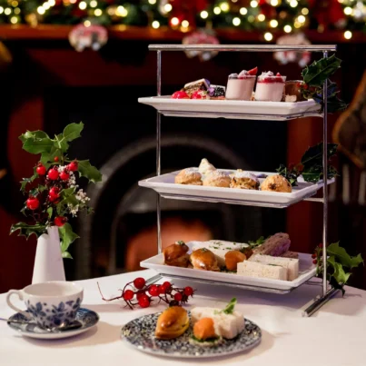 Festive three-tier afternoon tea stand with sandwiches, scones, mini desserts and holly by a cosy decorated fireplace