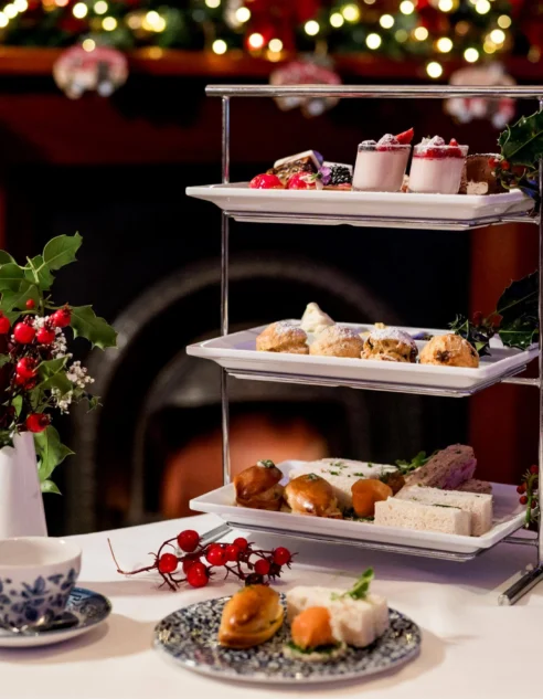 Festive three-tier afternoon tea stand with sandwiches, scones, mini desserts and holly by a cosy decorated fireplace